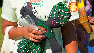 Pessoa vestindo camisa do projeto Quipea segura boneca de pano com roupas verdes estampadas durante evento cultural ao ar livre.