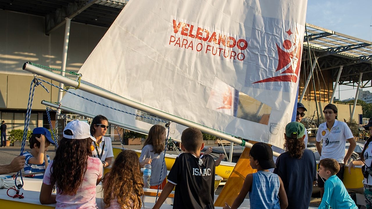 Um grupo de crianças observa um barco à vela amarelo enquanto um instrutor explica algo ao lado. No veleiro, há uma vela branca com o logotipo e a frase “Velejando para o Futuro”. A cena acontece ao ar livre, em um dia ensolarado, e as crianças usam roupas casuais e bonés.