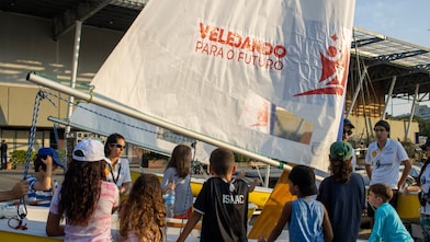Um grupo de crianças observa um barco à vela amarelo enquanto um instrutor explica algo ao lado. No veleiro, há uma vela branca com o logotipo e a frase “Velejando para o Futuro”. A cena acontece ao ar livre, em um dia ensolarado, e as crianças usam roupas casuais e bonés.
