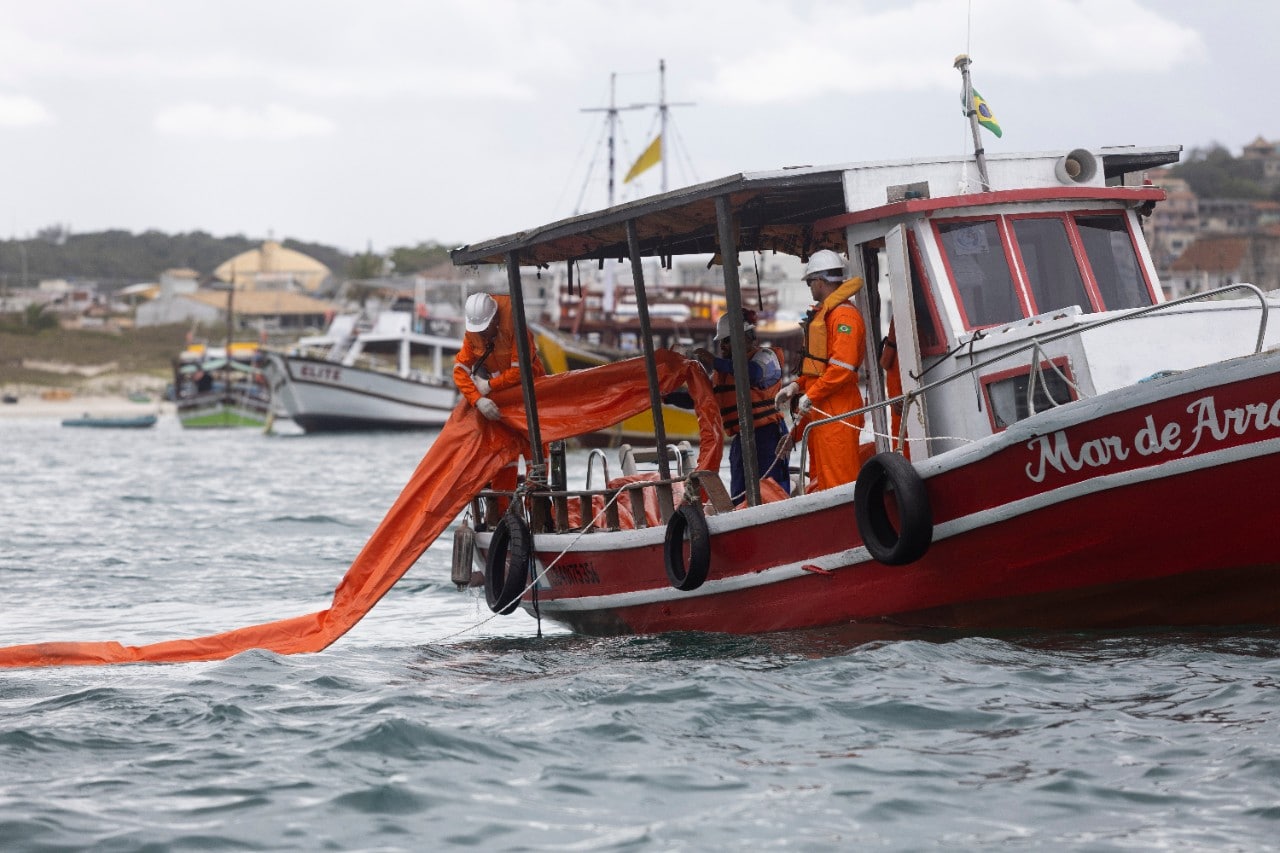 Profissionais usando macacões de segurança laranja, coletes salva-vidas e capacetes trabalham a bordo de uma embarcação vermelha, posicionando uma barreira flutuante laranja sobre a água. Ao fundo, outras embarcações estão ancoradas próximas à costa, sob céu nublado. A cena retrata uma operação marítima realizada em área costeira.