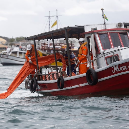 Profissionais usando macacões de segurança laranja, coletes salva-vidas e capacetes trabalham a bordo de uma embarcação vermelha, posicionando uma barreira flutuante laranja sobre a água. Ao fundo, outras embarcações estão ancoradas próximas à costa, sob céu nublado. A cena retrata uma operação marítima realizada em área costeira.