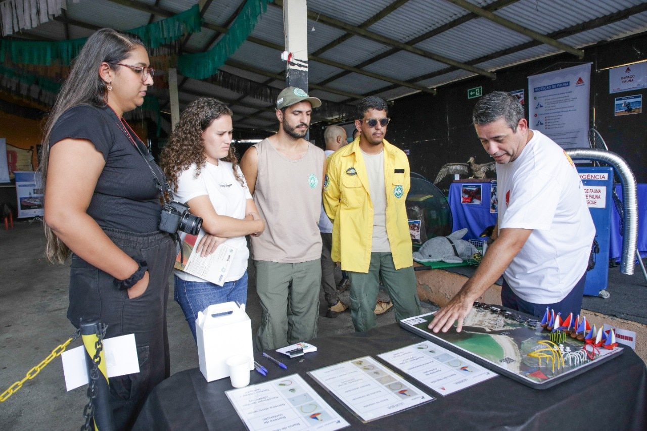 Grupo de cinco pessoas acompanha uma apresentação em um espaço coberto. Um homem aponta para um mapa e materiais informativos dispostos sobre uma mesa, enquanto os demais observam atentamente. Entre os participantes há profissionais de diferentes áreas, alguns com uniforme e outros com roupas casuais. Ao fundo, painéis educativos e materiais relacionados à fauna e ao meio ambiente compõem o ambiente expositivo.