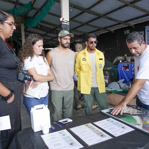 Grupo de cinco pessoas acompanha uma apresentação em um espaço coberto. Um homem aponta para um mapa e materiais informativos dispostos sobre uma mesa, enquanto os demais observam atentamente. Entre os participantes há profissionais de diferentes áreas, alguns com uniforme e outros com roupas casuais. Ao fundo, painéis educativos e materiais relacionados à fauna e ao meio ambiente compõem o ambiente expositivo.
