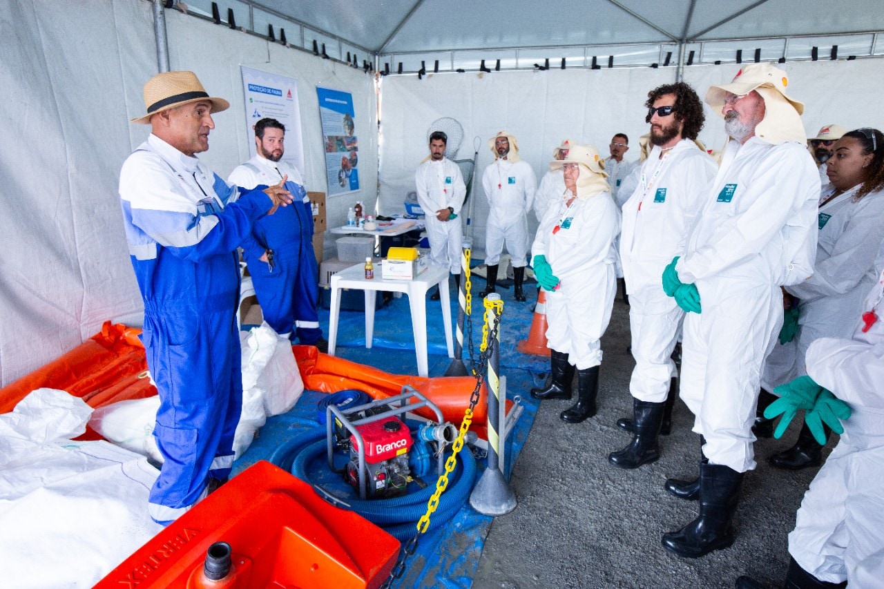 Interior de uma tenda usada para treinamento, onde um instrutor de chapéu bege e macacão azul fala a um grupo de pessoas vestidas com macacões brancos de proteção, luvas verdes e botas pretas. Alguns usam chapéus de aba larga e óculos de segurança. No primeiro plano, há barreiras laranja de contenção, uma bomba portátil com a marca 'Branco', mangueiras e outros equipamentos de resposta a derramamentos. Nas paredes da tenda, cartazes e materiais informativos reforçam o caráter instrucional do ambiente.