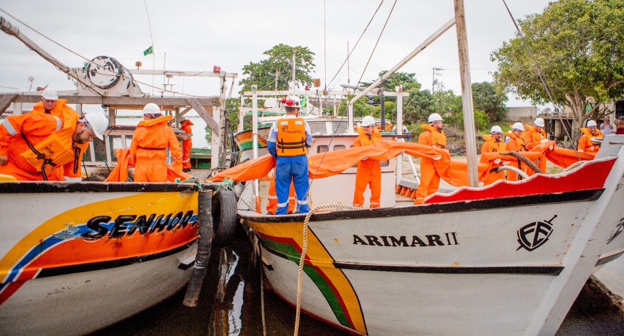 Equipe de profissionais usando macacões, coletes salva-vidas e capacetes laranja realiza uma operação entre duas embarcações atracadas. Os participantes manuseiam uma barreira flutuante laranja, estendida entre os barcos, em uma área portuária cercada por outras embarcações e vegetação. A atividade ocorre em ambiente aquático calmo, durante o dia.