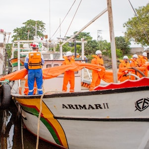 Equipe de profissionais usando macacões, coletes salva-vidas e capacetes laranja realiza uma operação entre duas embarcações atracadas. Os participantes manuseiam uma barreira flutuante laranja, estendida entre os barcos, em uma área portuária cercada por outras embarcações e vegetação. A atividade ocorre em ambiente aquático calmo, durante o dia.