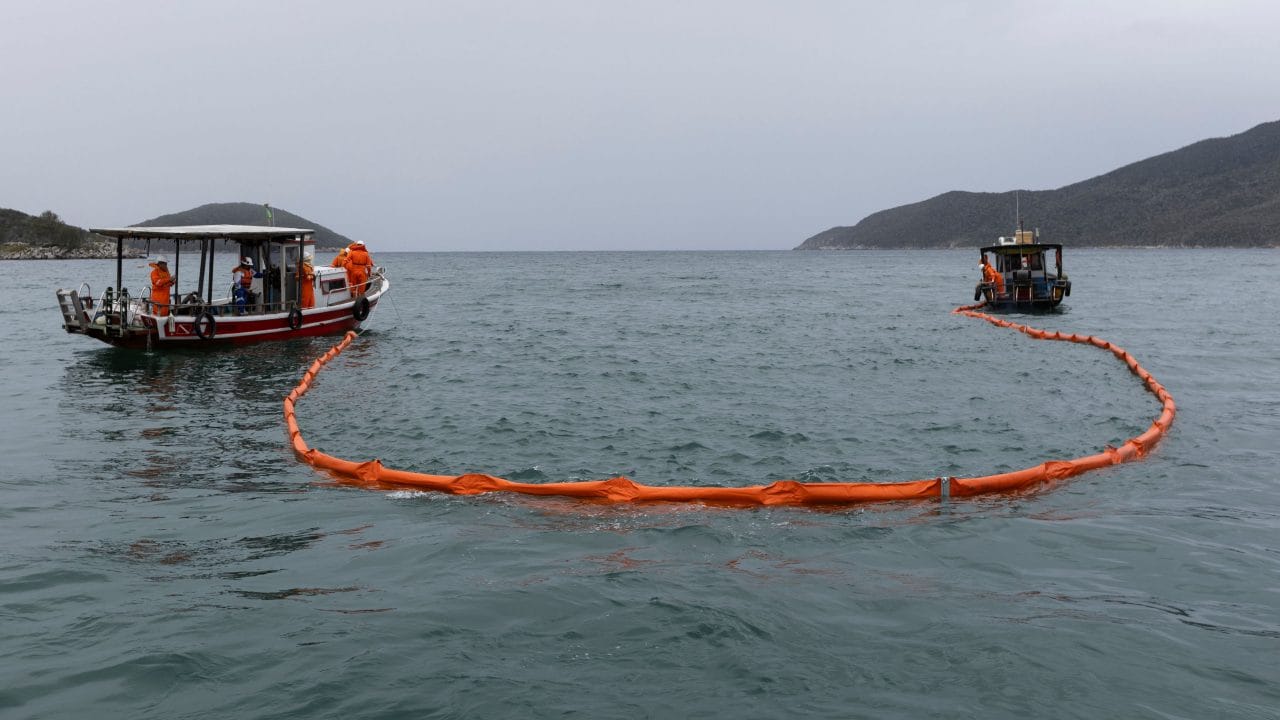 Duas embarcações de pequeno porte operam em mar aberto sob céu nublado. Entre elas, uma barreira flutuante laranja forma um grande círculo sobre a superfície da água. Profissionais usando equipamentos de segurança laranja trabalham a bordo dos barcos, enquanto ilhas e morros aparecem ao fundo, no horizonte. A cena retrata uma atividade de contenção ou monitoramento em ambiente marítimo.