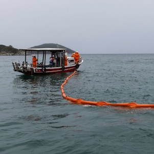 Duas embarcações de pequeno porte operam em mar aberto sob céu nublado. Entre elas, uma barreira flutuante laranja forma um grande círculo sobre a superfície da água. Profissionais usando equipamentos de segurança laranja trabalham a bordo dos barcos, enquanto ilhas e morros aparecem ao fundo, no horizonte. A cena retrata uma atividade de contenção ou monitoramento em ambiente marítimo.