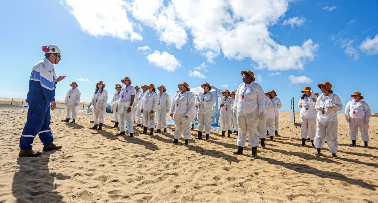 Grupo de profissionais reunidos em uma praia participa de uma atividade de orientação ao ar livre. Os participantes usam macacões brancos de proteção, chapéus, luvas e botas, enquanto um instrutor, vestido com uniforme azul e capacete, fala ao grupo. Ao fundo, há uma tenda de apoio e equipamentos de campo. O céu azul com nuvens e a faixa de areia iluminada pelo sol compõem a cena.