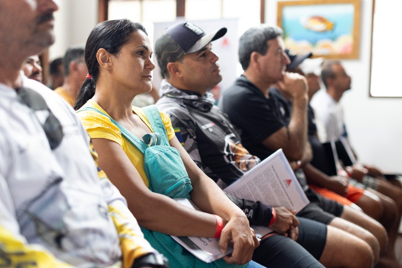 Grupo de pessoas sentadas em fileiras dentro de um espaço iluminado por luz natural, segurando cadernos e materiais impressos enquanto acompanham atentamente uma apresentação ou palestra. O ambiente sugere um encontro formal, como um seminário ou oficina, com um quadro emoldurado pendurado na parede ao fundo.