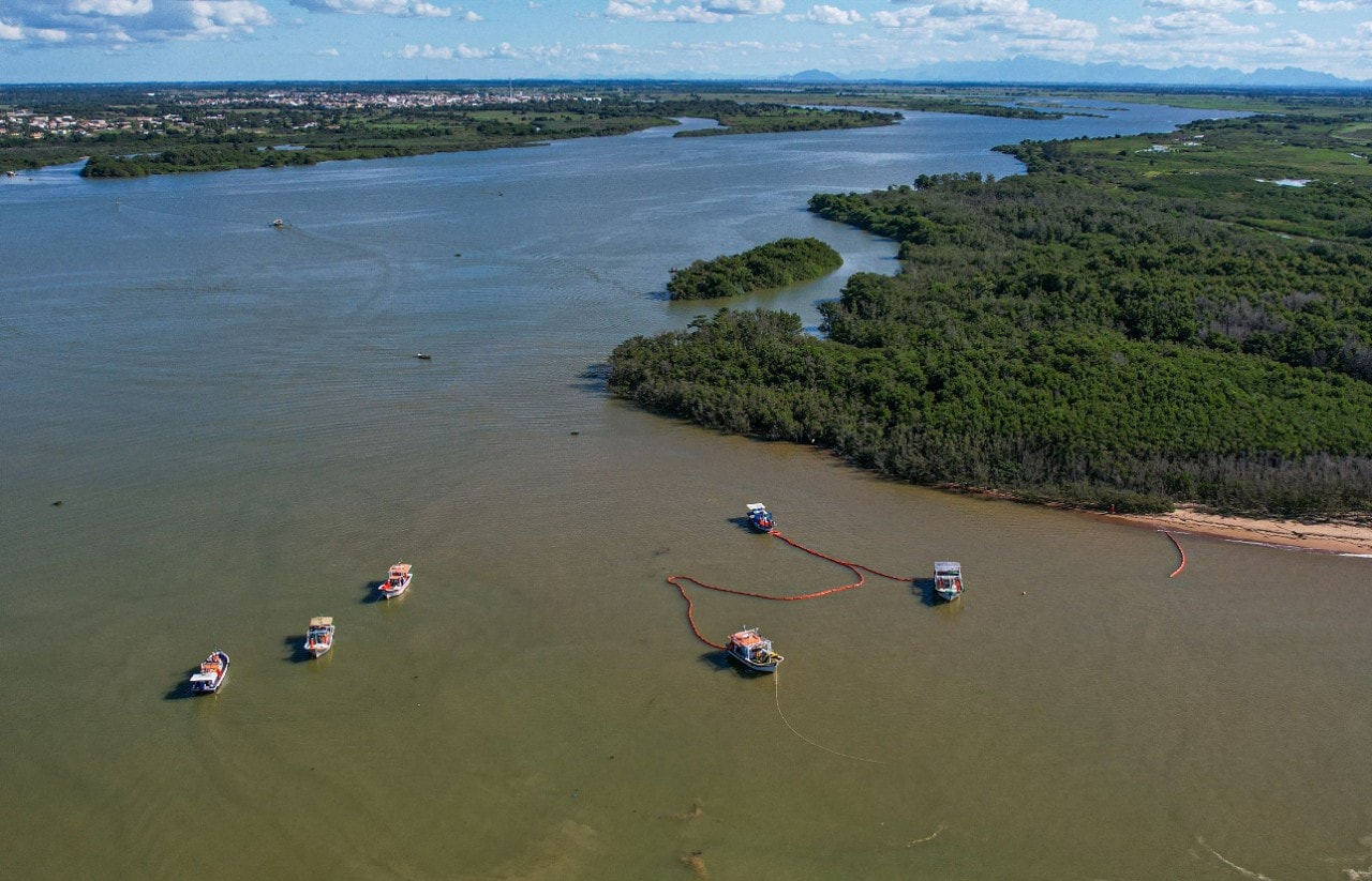 Vista aérea de uma ampla área estuarina cercada por manguezais e vegetação costeira. Seis embarcações de pequeno porte estão distribuídas sobre a água, algumas conectadas por barreiras flutuantes laranjas posicionadas próximas à margem. Ao fundo, o curso d’água se estende em direção ao horizonte, onde são visíveis áreas urbanas, campos e formações montanhosas distantes. A cena retrata uma operação marítima em ambiente costeiro.