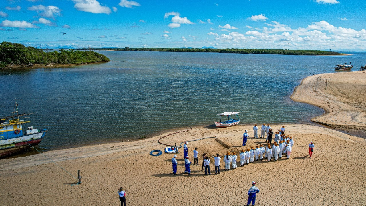 Praia de areia clara ao lado de um corpo de água calmo, com barcos ancorados próximos à margem — incluindo um azul e amarelo à esquerda e um branco com cobertura ao centro. Um grupo de pessoas em trajes de proteção brancos e uniformes azuis está reunido sobre a areia, participando de uma atividade organizada. O céu azul com nuvens esparsas e a vegetação verde na margem oposta completam o cenário.
