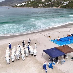 Vista aérea de uma praia cercada por mar azul-esverdeado e morros ao fundo. Na faixa de areia, uma equipe de profissionais vestindo macacões brancos de proteção, chapéus e botas participa de uma atividade de orientação em grupo. Próximo a eles, há tendas de apoio, equipamentos organizados sobre lonas azuis e áreas delimitadas por cordas. Embarcações podem ser vistas ao longe, próximas à costa.