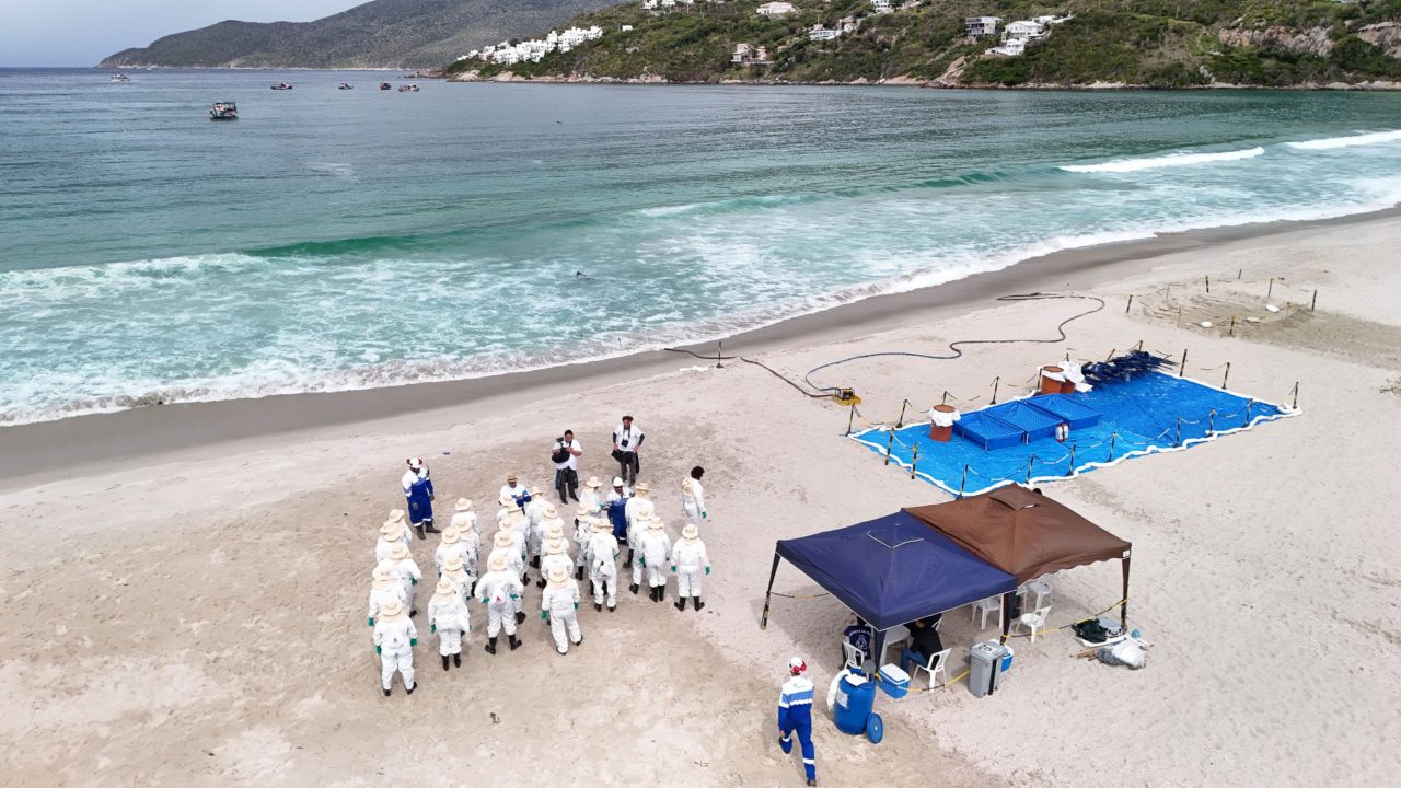 Grupo de cerca de 40 pessoas posa para uma foto em frente a tendas montadas em uma área externa. Os participantes usam diferentes tipos de vestimenta, incluindo uniformes, macacões de proteção e roupas casuais, e alguns seguram certificados. Ao fundo, uma faixa com a inscrição “Exercício Simulado” está afixada na estrutura principal. A cena registra o encerramento ou a celebração de uma atividade coletiva de treinamento, com edifícios e veículos ao redor.