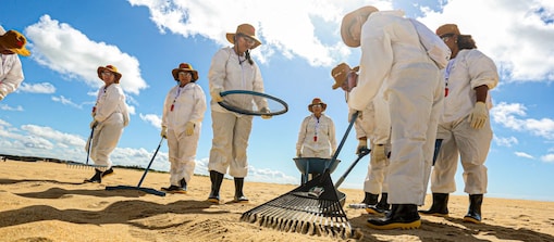 Grupo de profissionais utiliza equipamentos de limpeza e peneiramento em uma faixa de areia sob céu azul com nuvens. Vestidos com macacões brancos de proteção, luvas, botas e chapéus, os participantes realizam atividades de inspeção e coleta na praia. A imagem é registrada em ângulo baixo, destacando a equipe reunida ao redor da área de trabalho.
