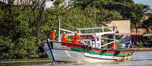 Embarcação de pequeno porte navega por um canal cercado por vegetação às margens. A bordo, profissionais usando macacões de segurança laranja e capacetes realizam atividades operacionais. O barco branco, verde e vermelho segue sobre águas calmas, com construções e outras embarcações visíveis ao fundo.