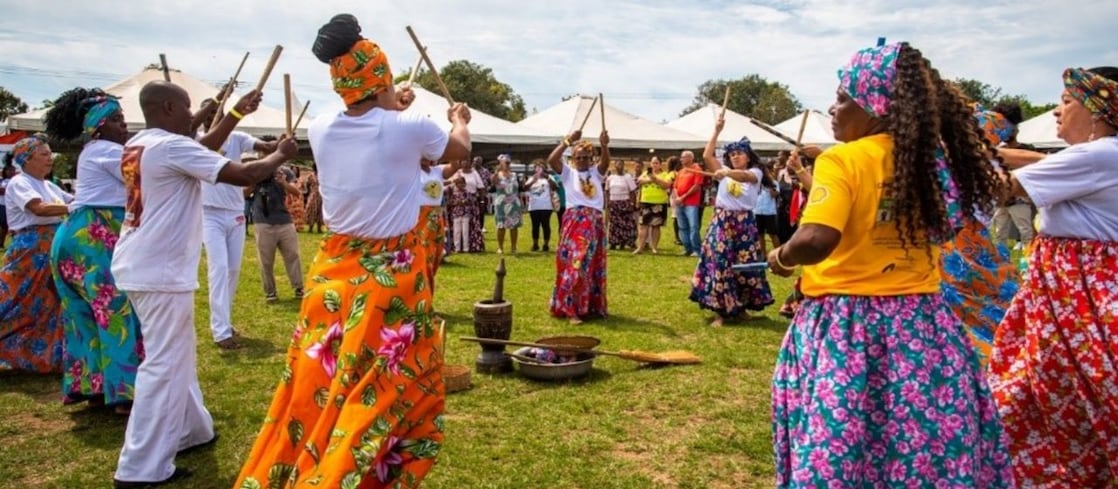 Homens e mulheres negros formam uma roda festiva em um grande gramado, batendo bastões em madeira ritmicamente, ao alto da cabeça. Elas vestem saias floridas e turbantes coloridos, eles vestem camiseta e calça brancas, e se apresentam para um público ao seu redor.
