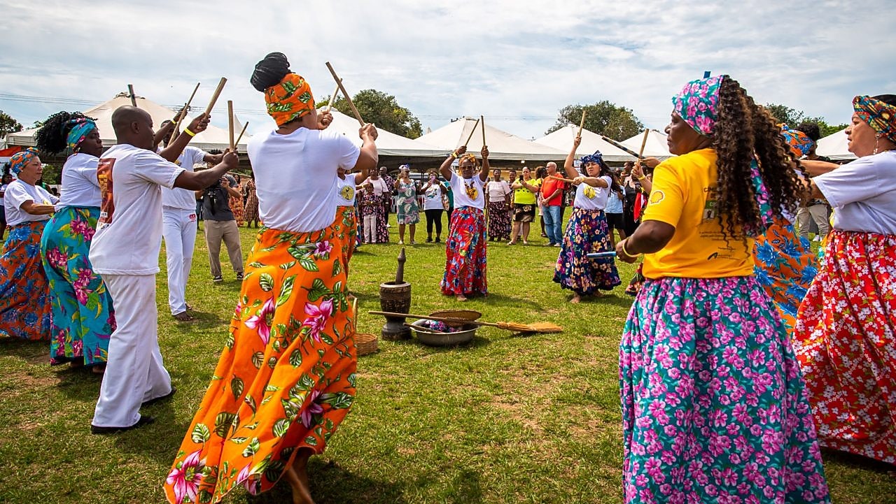 Grupo de mulheres, vestidas com saias longas floridas, dançando maculelê.