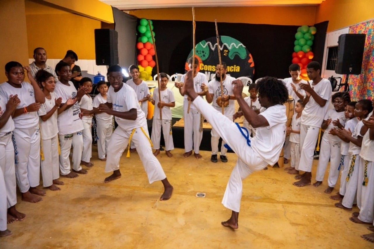 Roda de capoeira com jovens e crianças negras vestidos de branco com cordas amarradas à cintura. Dois capoeiristas jogam no centro, enquanto os demais batem palmas e tocam instrumentos tradicionais (como berimbau e atabaque). O ambiente é de energia, cultura e ancestralidade.