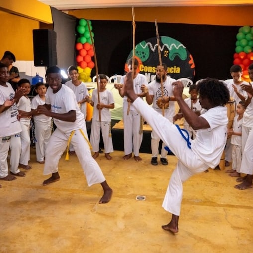 Roda de capoeira com jovens e crianças negras vestidos de branco com cordas amarradas à cintura. Dois capoeiristas jogam no centro, enquanto os demais batem palmas e tocam instrumentos tradicionais (como berimbau e atabaque). O ambiente é de energia, cultura e ancestralidade.
