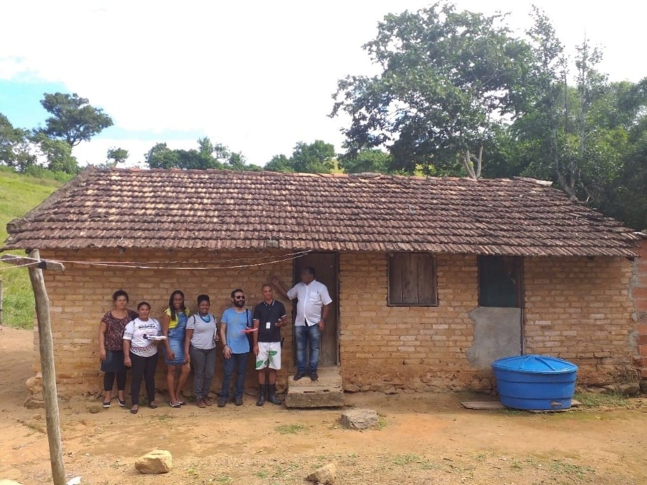 Um grupo de pessoas está reunido em frente a uma casa simples de tijolos e telhado de barro. O local parece ser uma área rural, com vegetação ao fundo. Todos estão alinhados sorrindo para a foto, sugerindo um momento de visita ou registro de memória.