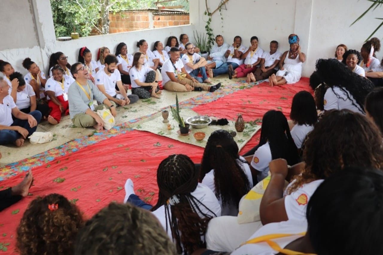 Um grupo grande de pessoas está reunido em círculo dentro de um espaço coberto. Elas estão sentadas no chão, algumas sobre esteiras de palha, e vestem camiseta branca. No centro do círculo há objetos de cerâmica, folhas e plantas sobre um pano vermelho. formando um cenário ritualístico. À direita, uma mulher veste roupa branca e turbante azul e vermelho, gesticulando enquanto fala. As demais pessoas observam com atenção.