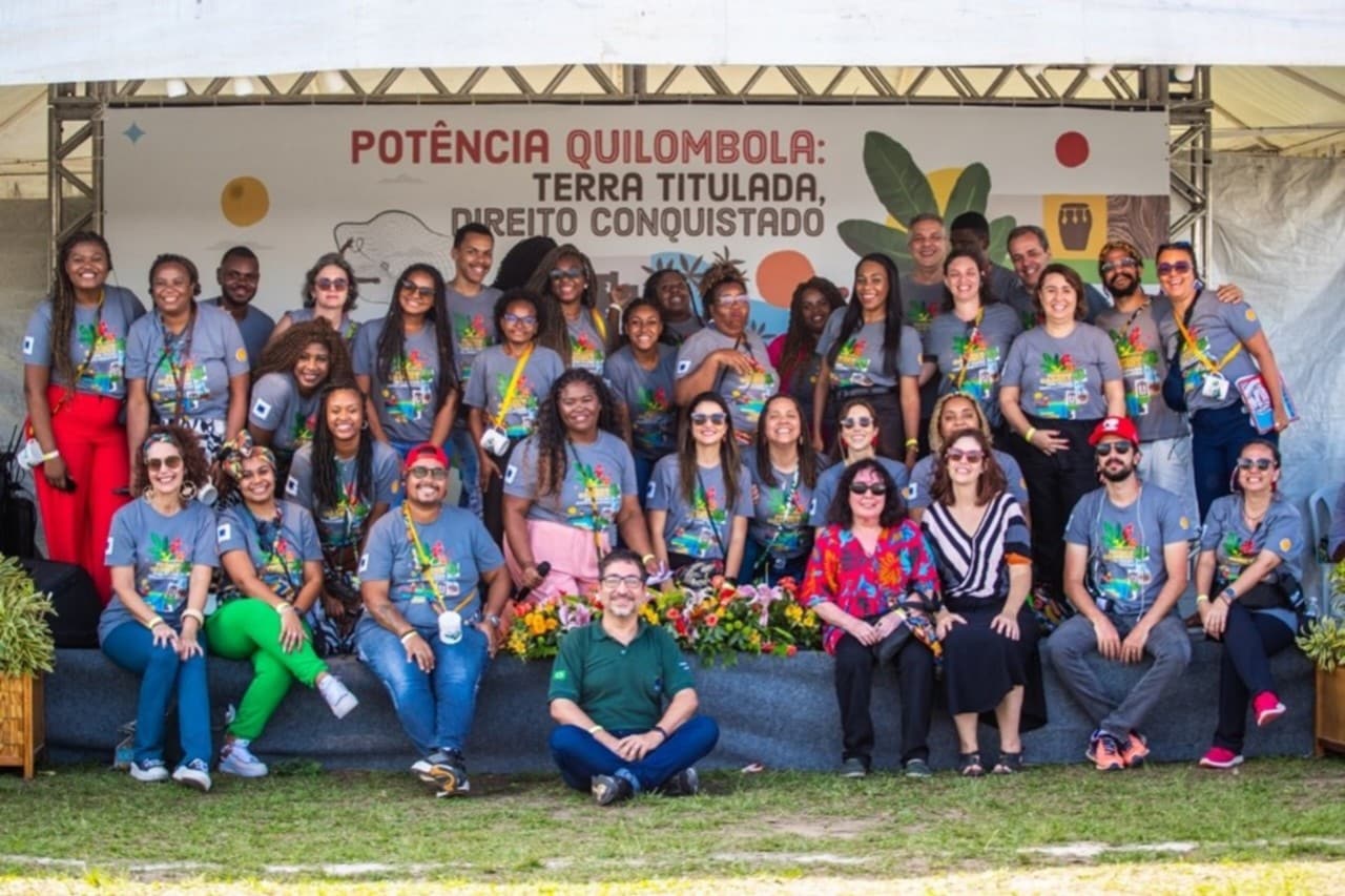 Foto coletiva com cerca de 30 pessoas sorridentes, reunidas em frente a um painel com a frase: “Potência Quilombola: Terra Titulada, Direito Conquistado”.
