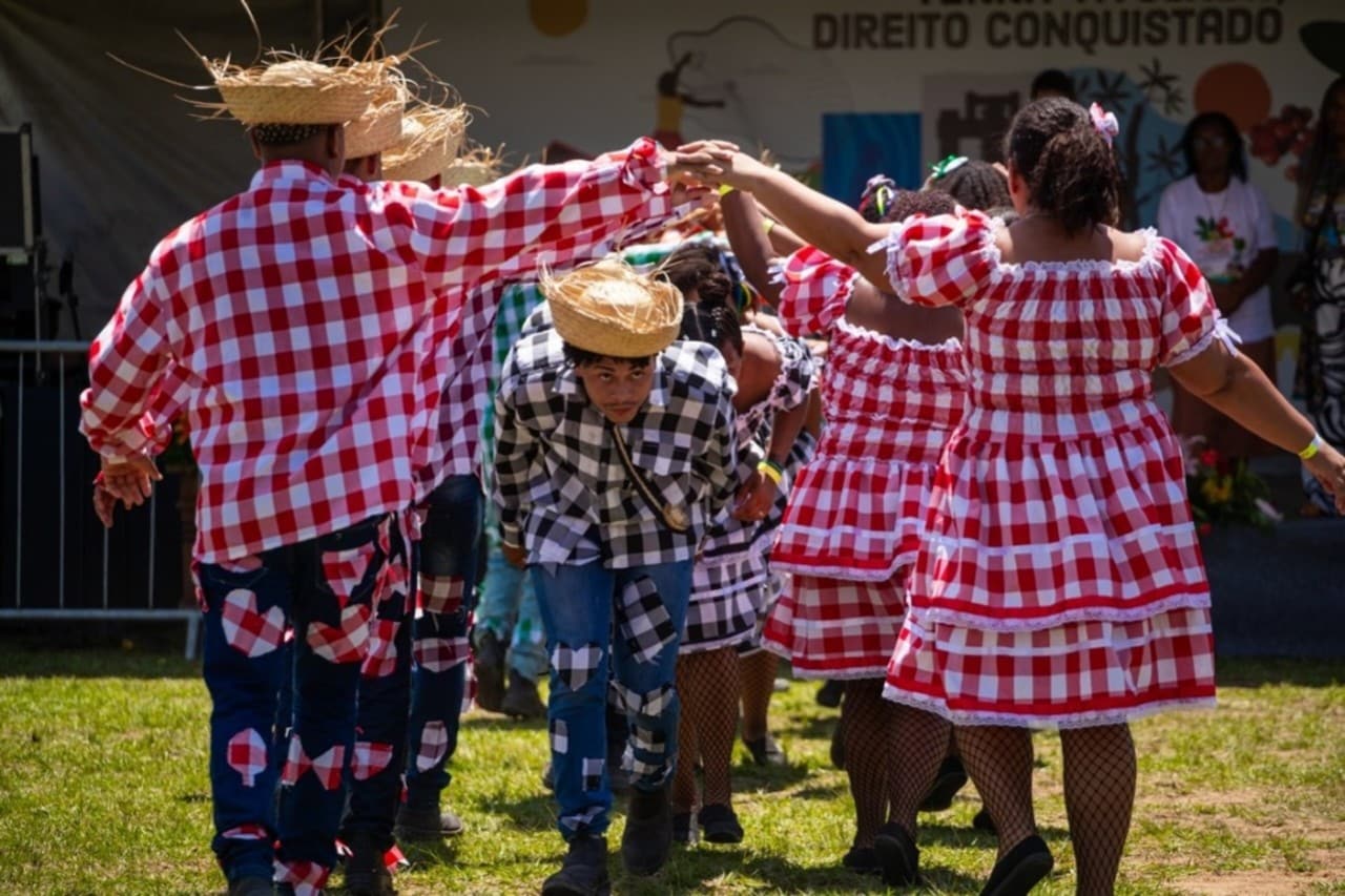 Um grupo de pessoas, homens e mulheres, participa de uma dança típica, uma quadrilha. Eles vestem roupas tradicionais com estampas de quadrados coloridos (xadrez vermelho e branco ou preto e branco) e chapéus de palha. Estão formando um túnel com os braços erguidos, por onde outros participantes passam, uma cena típica de festa junina brasileira.