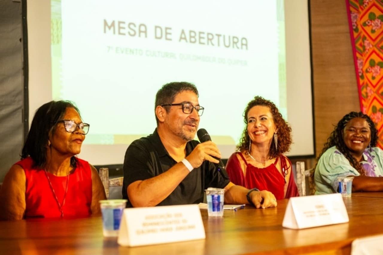 Uma mesa de abertura de evento. Quatro pessoas, sendo duas mulheres negras na ponta, um homem e uma mulher branca ao meio, sentadas à frente de um painel projetado que diz: “Mesa de Abertura – 7º Evento Cultural Quilombola do Quipea”. Todos estão sorrindo.