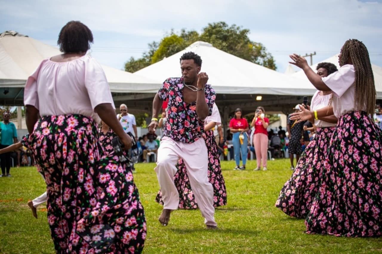 Grupo de Ciranda apresenta em uma coreografia no centro de um gramado verde. Ao fundo estão posicionadas duas grandes barracas de lona branca. Ao centro de uma roda, um homem negro dança com expressão intensa, e mulheres negras ao redor batem palmas. Elas vestem saias floridas compridas e blusas brancas. Ele veste calça branca e blusa florida, com a mesma estampa das saias das mulheres.
