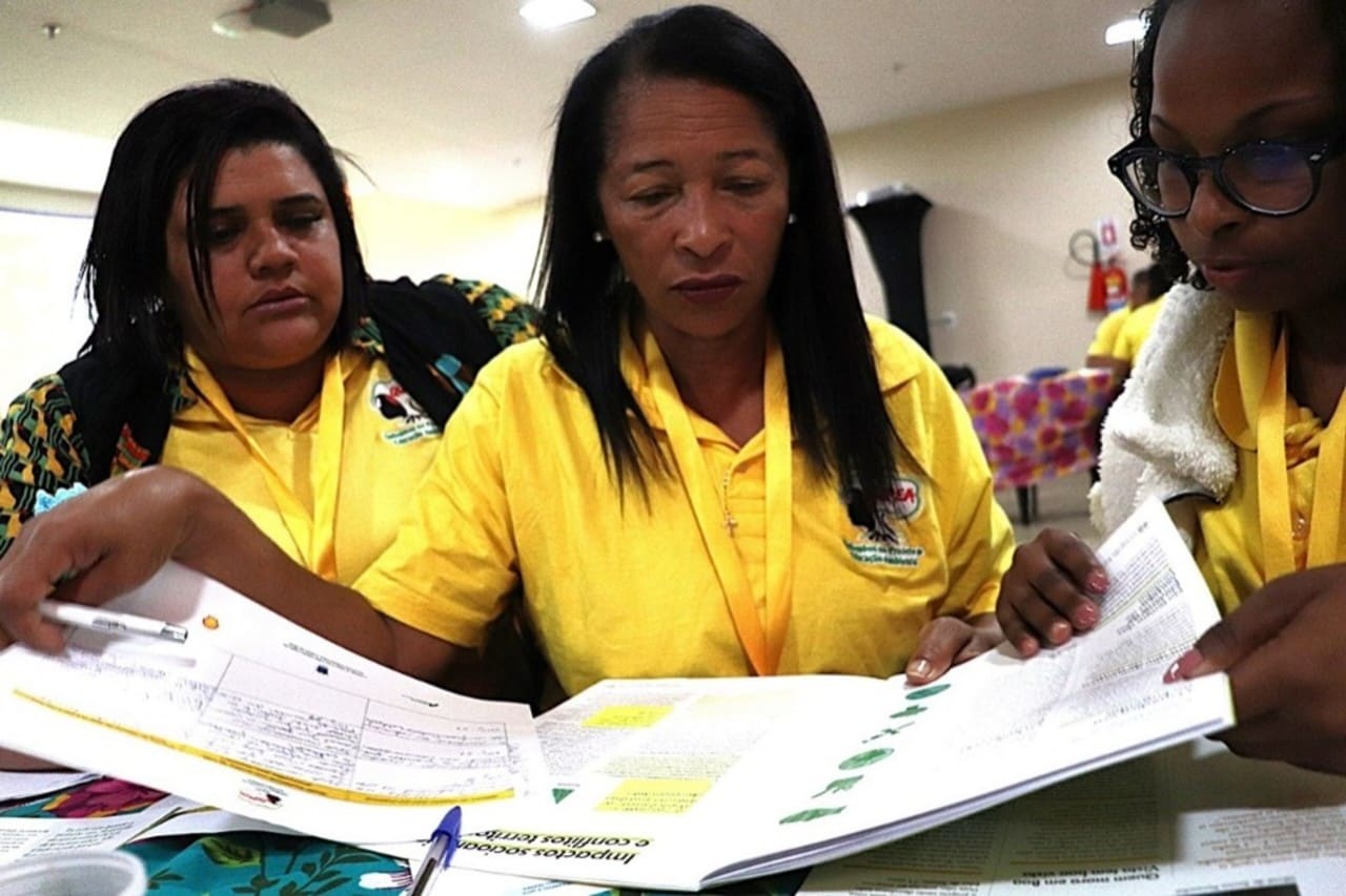 Três mulheres negras examinam de forma concentrada uma revista e alguns papéis, em momento de estudo coletivo. As três vestem camiseta amarela com o logotipo do Quipea à esquerda do peito, e portam crachás com fita amarela no pescoço.
