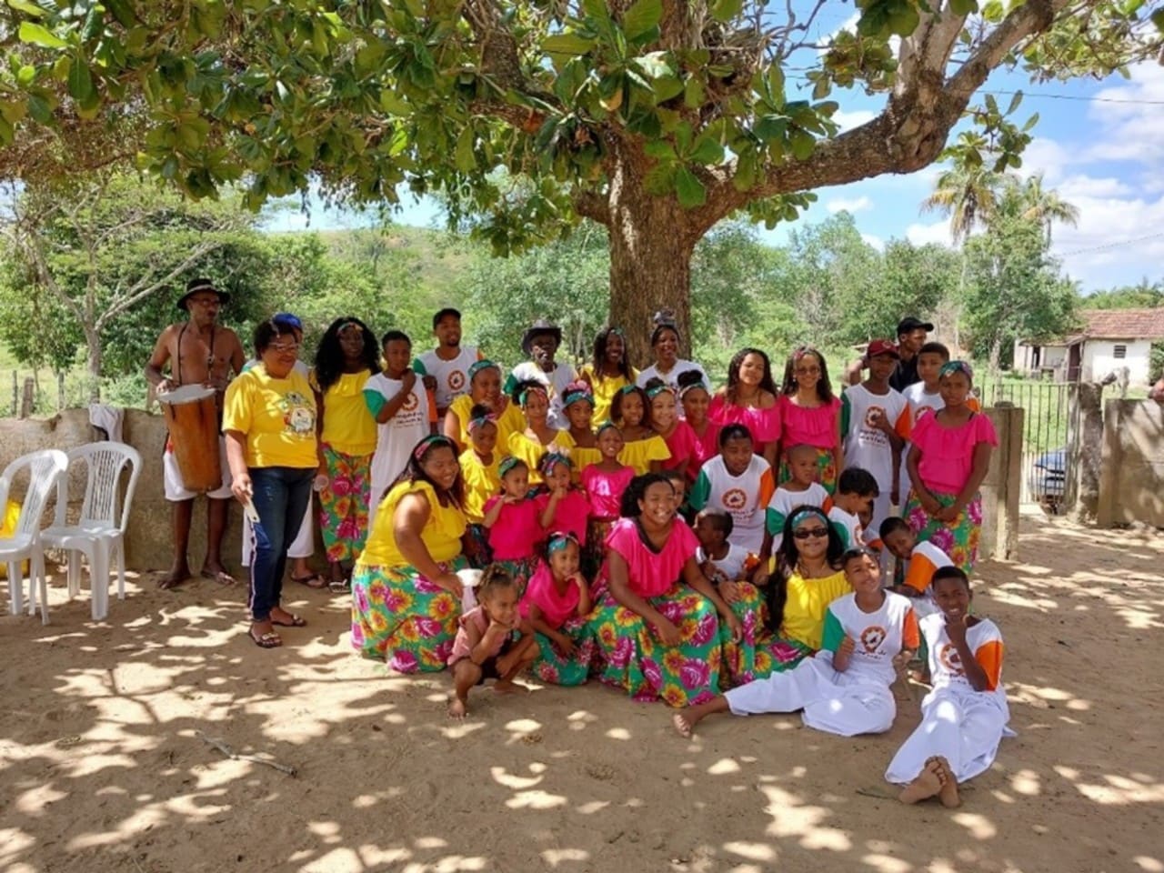 Um grande grupo de pessoas, incluindo crianças, jovens e adultos negros, está reunido sob uma árvore. Eles vestem roupas coloridas, muitas mulheres e meninas com saias estampadas e blusas amarelas ou rosas. Algumas pessoas usam camisas com logotipos de associação quilombola. O clima é festivo e comunitário.