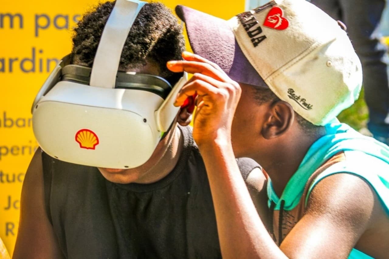 Dois jovens negros interagem com óculos de realidade virtual da Shell. Um deles usa o aparelho enquanto o outro observa de perto, demonstrando curiosidade. Eles estão em um ambiente iluminado, com um fundo parcialmente amarelo que parece ser parte de um painel em um evento.