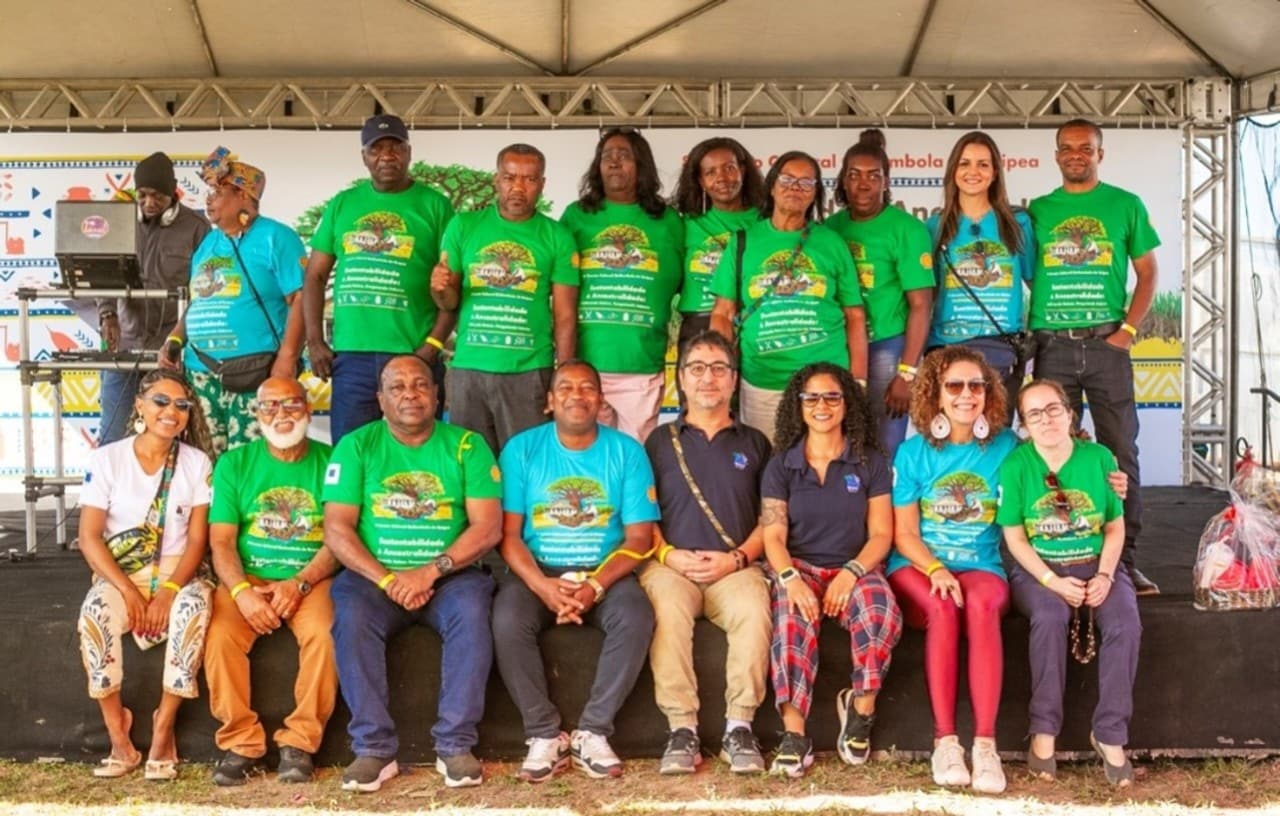 Grupo diverso de pessoas posa sorridente em um palco durante um evento cultural. A maioria veste camisetas verdes com o tema “Sustentabilidade & Ancestralidade”, sugerindo um encontro voltado à valorização da cultura quilombola ou afrodescendente. Ao fundo, há um banner decorado com grafismos que remetem ao cenário de uma comunidade quilombola tradicional.