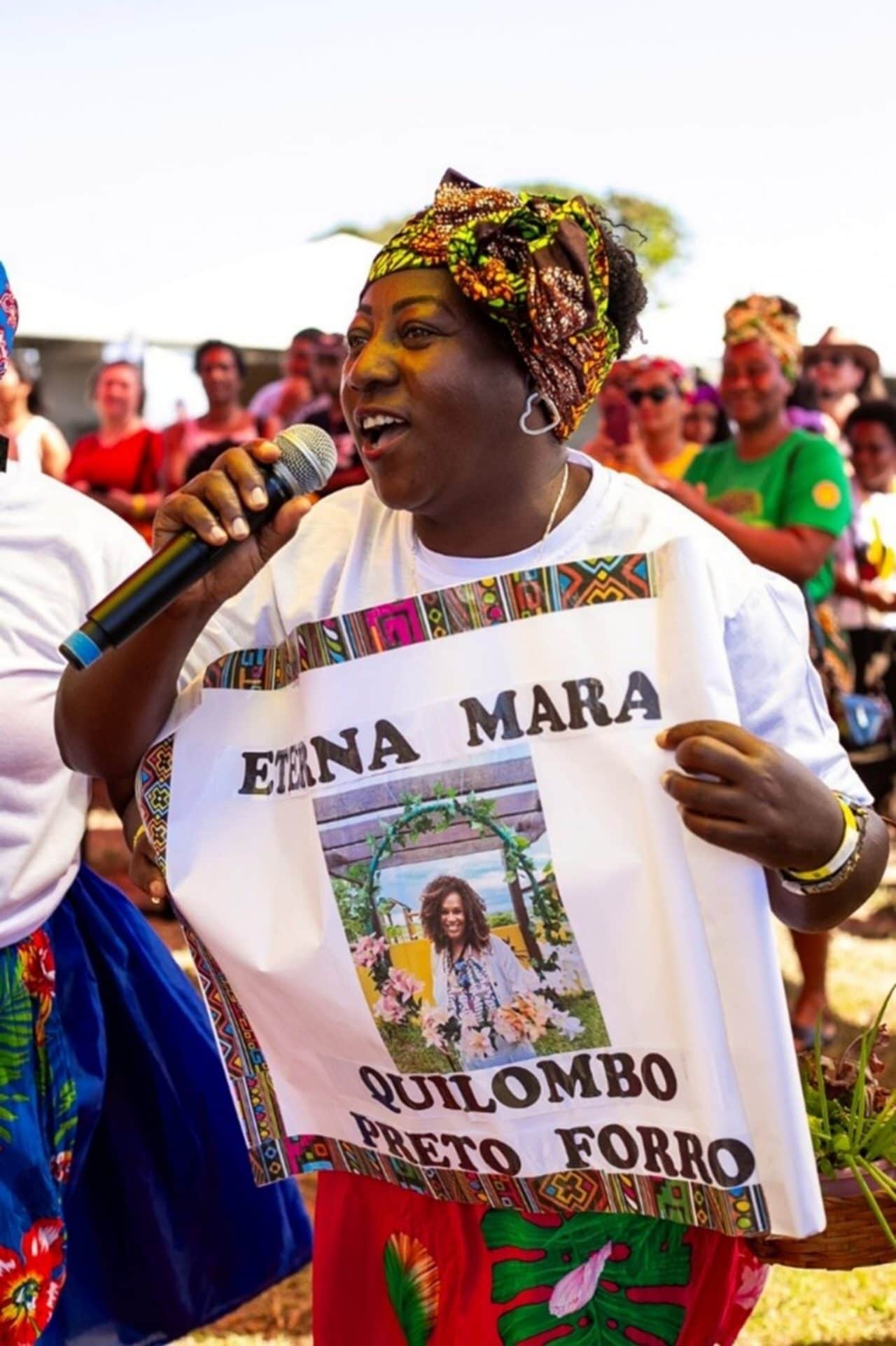 Uma mulher negra segura um microfone e fala ou canta. Ela veste uma camisa branca personalizada com a frase “Eterna Mara - Quilombo Preto Forro”, acompanhada da foto de uma mulher homenageada. Pessoas ao fundo acompanham. 
