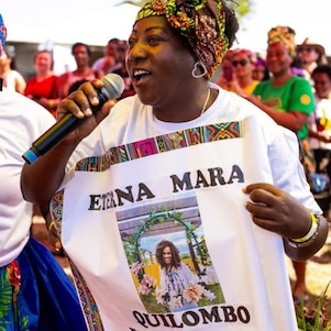 Uma mulher negra segura um microfone e fala ou canta. Ela veste uma camisa branca personalizada com a frase “Eterna Mara - Quilombo Preto Forro”, acompanhada da foto de uma mulher homenageada. Pessoas ao fundo acompanham.