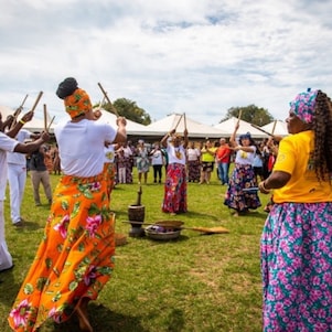 Homens e mulheres negros formam uma roda festiva em um grande gramado, batendo bastões em madeira ritmicamente, ao alto da cabeça. Elas vestem saias floridas e turbantes coloridos, eles vestem camiseta e calça brancas, e se apresentam para um público ao seu redor.