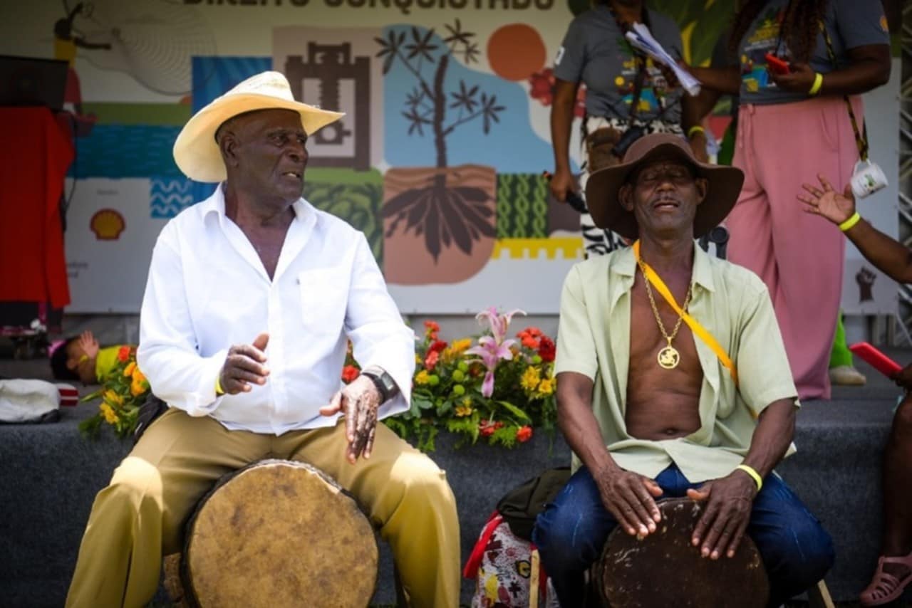 Dois homens negros sentados em cima de tambores, tocando. Um usa chapéu de palha e camisa branca, o outro está com camisa verde-claro aberta, e usa chapéu marrom e colar dourado. Ao fundo dos dois está um palco com painel colorido e flores.