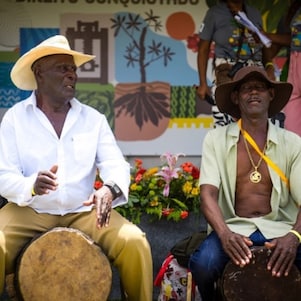 Dois homens negros sentados em cima de tambores, tocando. Um usa chapéu de palha e camisa branca, o outro está com camisa verde-claro aberta, e usa chapéu marrom e colar dourado. Ao fundo dos dois está um palco com painel colorido e flores.