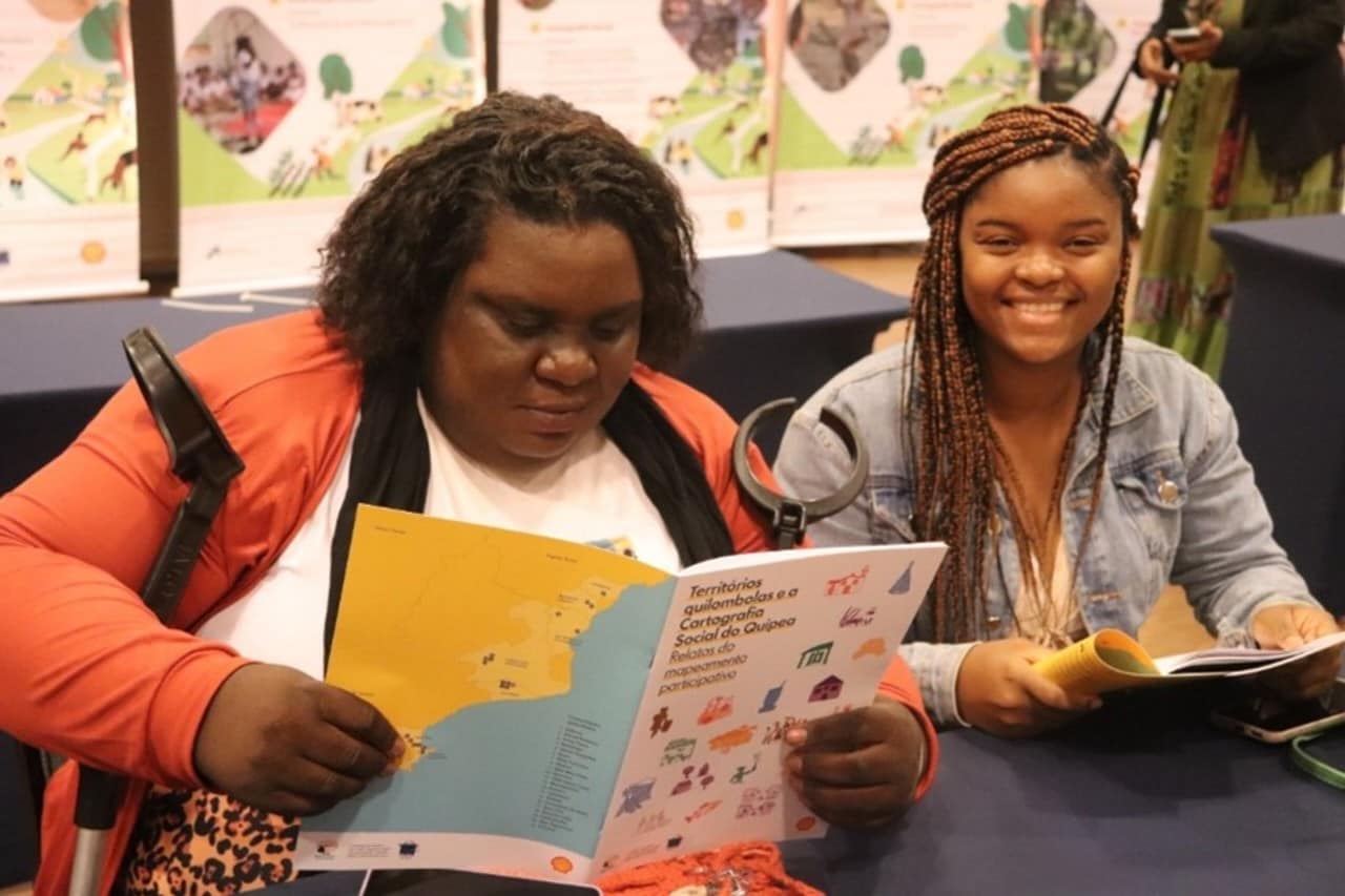 Duas mulheres negras manuseiam o 22º fascículo da Cartografia Social do Quipea, em formato de revista. Uma delas lê atentamente a revista aberta, e usa camiseta branca e casaco laranja. A outra sorri para a câmera, e usa jaqueta jeans e cabelo trançado, comprido.