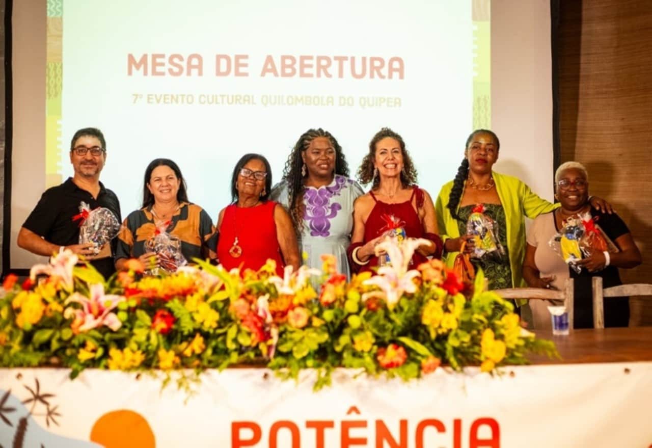 Grupo de sete pessoas em pé, atrás da mesa decorada com flores. No fundo, há a inscrição em uma tela grande “Mesa de Abertura – 7º Evento Cultural Quilombola do Quipea”. Elas seguram lembranças ou presentes recebidos.