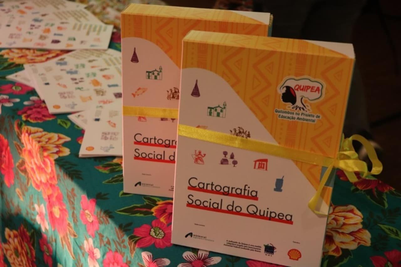Duas caixas personalizadas, decoradas com desenhos e o logotipo do Quipea e envoltas com fita de cetim amarela, onde se lê Cartografia Social do Quipea. Elas estão dispostas sobre uma toalha florida.