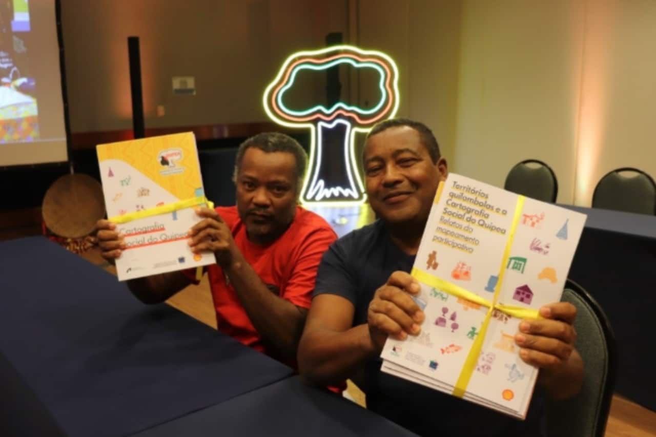 Dois homens negros estão sentados lado a lado em uma mesa azul. Eles sorriem e seguram, cada um, uma caixa dos 22 fascículos do projeto Quipea, sobre cartografia social quilombola. Atrás deles, há cadeiras pretas vazias e um display iluminado em formato de árvore colorida.