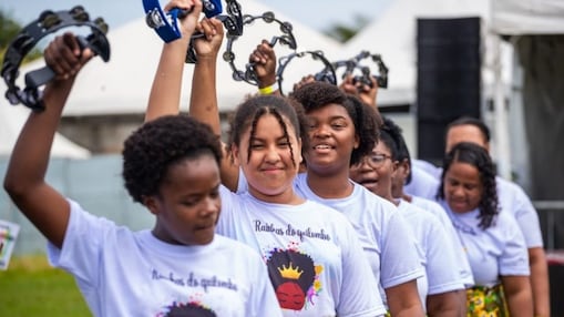 Grupo de mulheres negras participa de uma apresentação ao ar livre, vestindo camisetas brancas com o logo “Rainhas do Quimbanda” e saias coloridas com estampas africanas. Elas estão alinhadas em fila, sorrindo e levantando pandeiros no ar, em um momento de celebração e alegria.