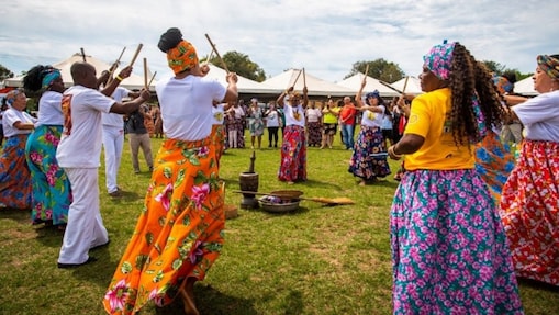 Homens e mulheres negros formam uma roda festiva em um grande gramado, batendo bastões em madeira ritmicamente, ao alto da cabeça. Elas vestem saias floridas e turbantes coloridos, eles vestem camiseta e calça brancas, e se apresentam para um público ao seu redor.