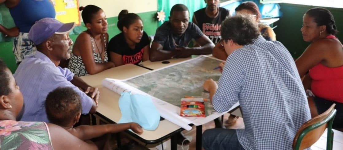 Reunião em uma sala de escola. Ao redor de uma mesa, moradores de comunidade quilombola observam atentamente um mapa de papel grande sobre uma mesa, enquanto um homem branco vestido com camisa xadrez azul e branca aponta para o centro mapa.
