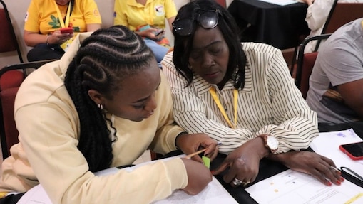 Duas mulheres negras estudando juntas sentadas lado a lado em uma mesa, analisam documentos impressos. Uma delas aponta para o texto, enquanto a outra segura um papel adesivo, indicando trabalho colaborativo e atenção. A mulher da esquerda veste moletom bege e tem cabelo trançado. A mulher da direita veste blusa listrada bege e marrom, e tem óculos de grau no alto da cabeça.