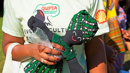 Detalhe da mão de uma pessoa negra segurando uma boneca de pano preta, vestida com tecido verde estampado e um turbante da mesma cor. A pessoa veste uma camiseta clara com o logotipo do projeto Quipea e o símbolo da Shell.