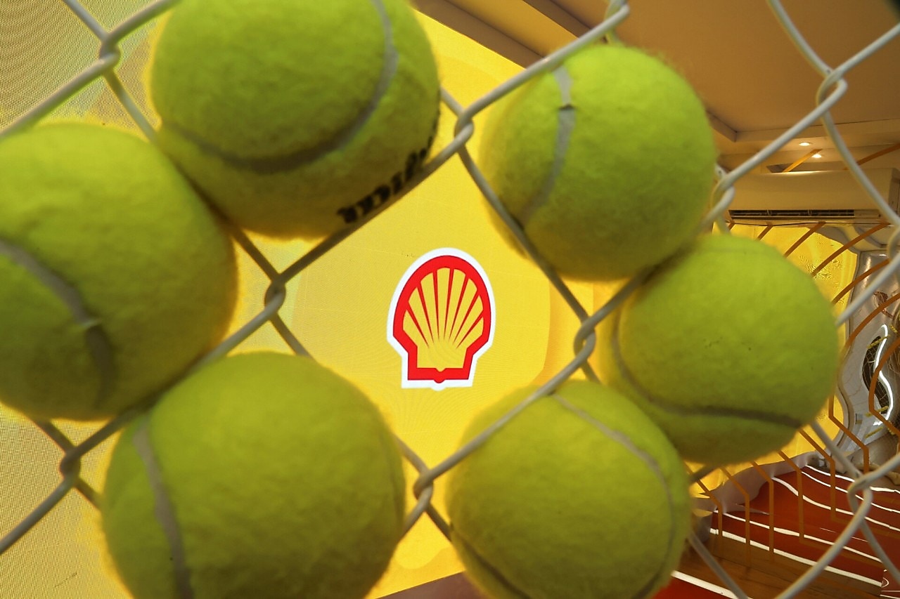 Bolas de tênis vistas através de uma grade metálica, com o logotipo da Shell ao fundo em um espaço decorado para evento esportivo.