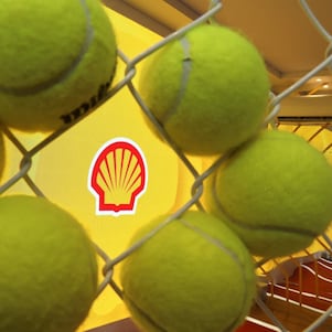 Bolas de tênis vistas através de uma grade metálica, com o logotipo da Shell ao fundo em um espaço decorado para evento esportivo.